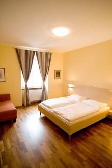 Útulný dvoulůžkový pokoj s velkou postelí, dřevěnou podlahou, křeslem a oknem se závěsy.