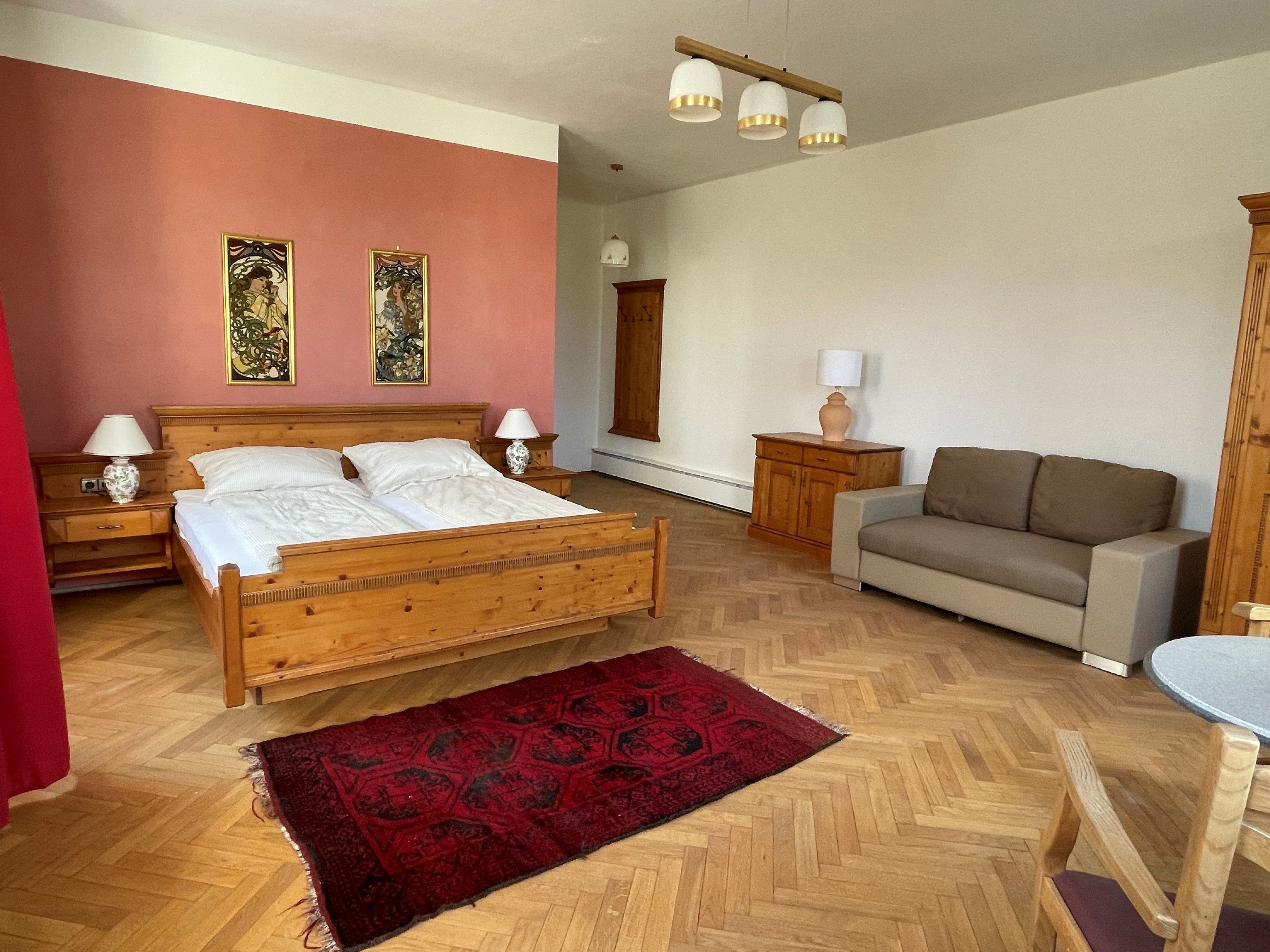 Prostorná ložnice s dřevěnou postelí, nočními stolky, pohovkou a červeným kobercem na dřevěné podlaze.