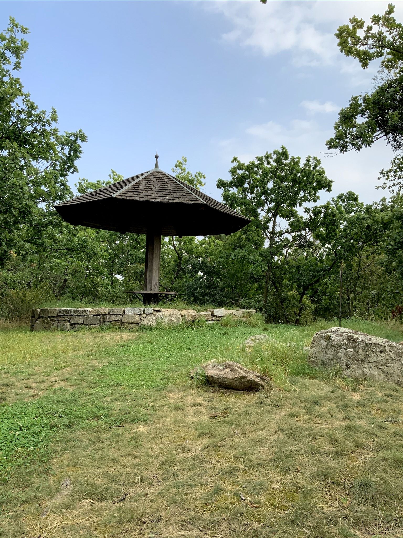 Dřevěný pavilon se špičatou střechou v zeleném lesním prostředí.