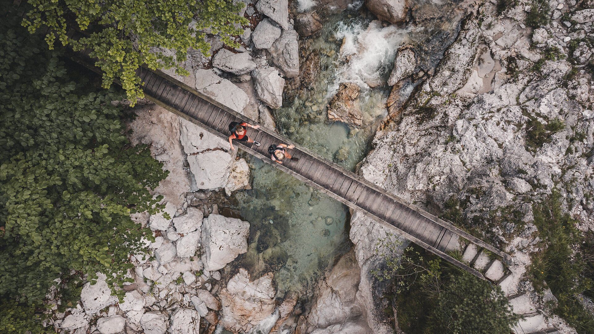 Zwei Wanderinnen gehen über eine schmale Holzbrücke, die über einen klaren, von Felsen umgebenen Fluss in einer Schlucht führt