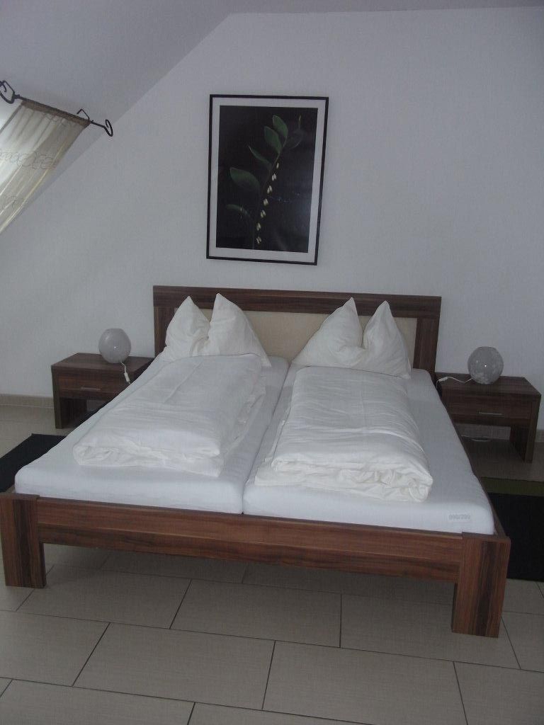 Manželská postel v hotelovém pokoji s bílým ložním prádlem a obrázkem rostliny na stěně.