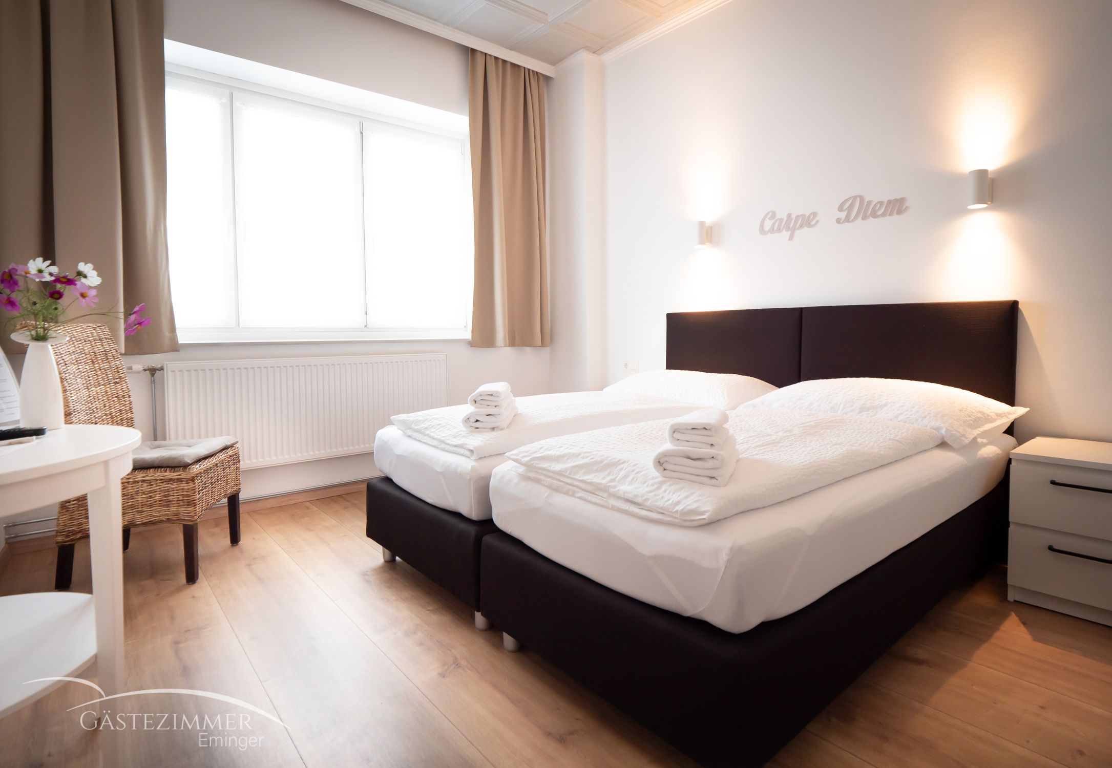 Moderní dvoulůžkový pokoj se dvěma postelemi, bílými přikrývkami a ručníky. Nápis "Carpe Diem" na stěně, světlé závěsy a dřevěná podlaha.