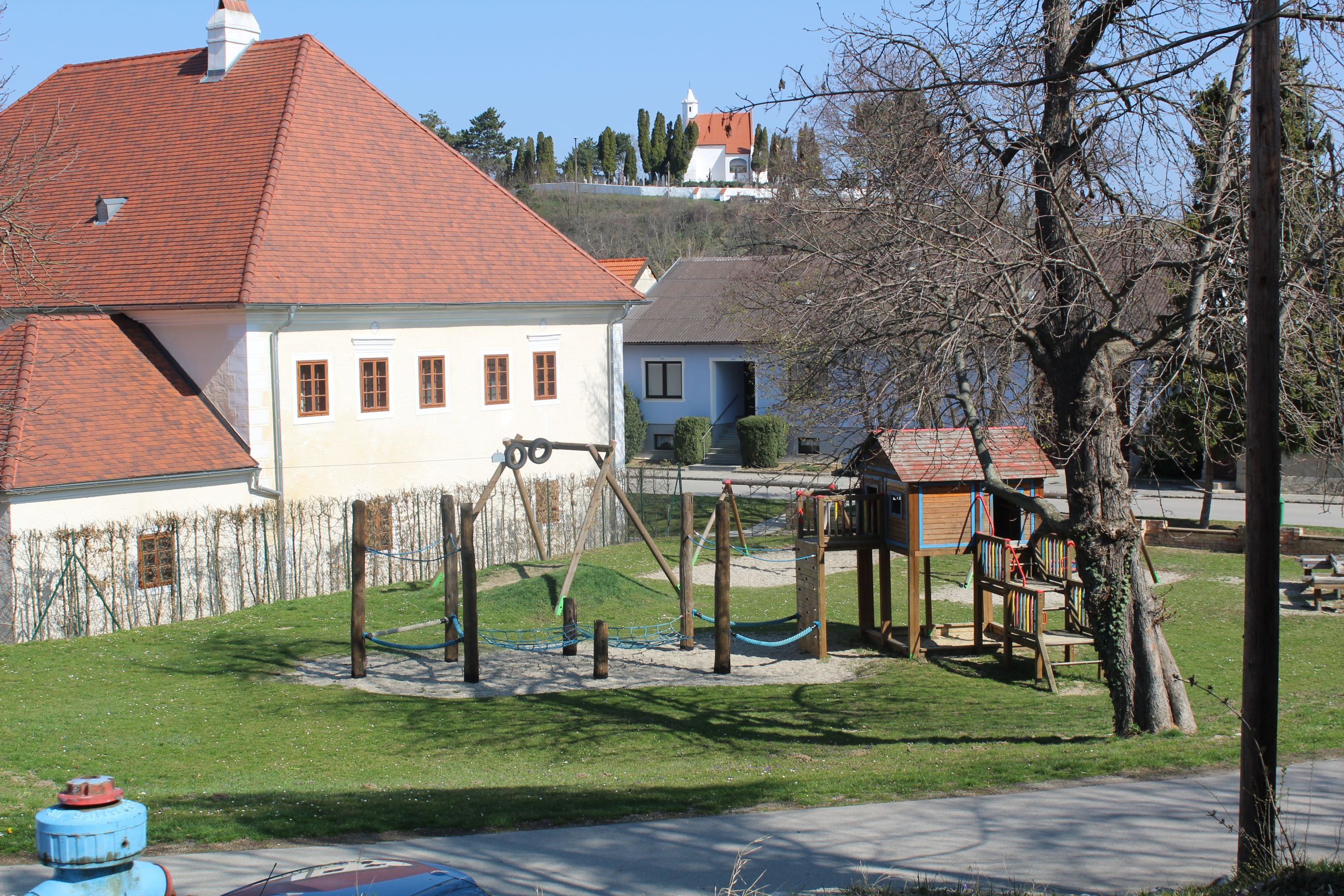 Dětské hřiště s prolézačkami a houpačkami vedle budovy s červenou střechou.