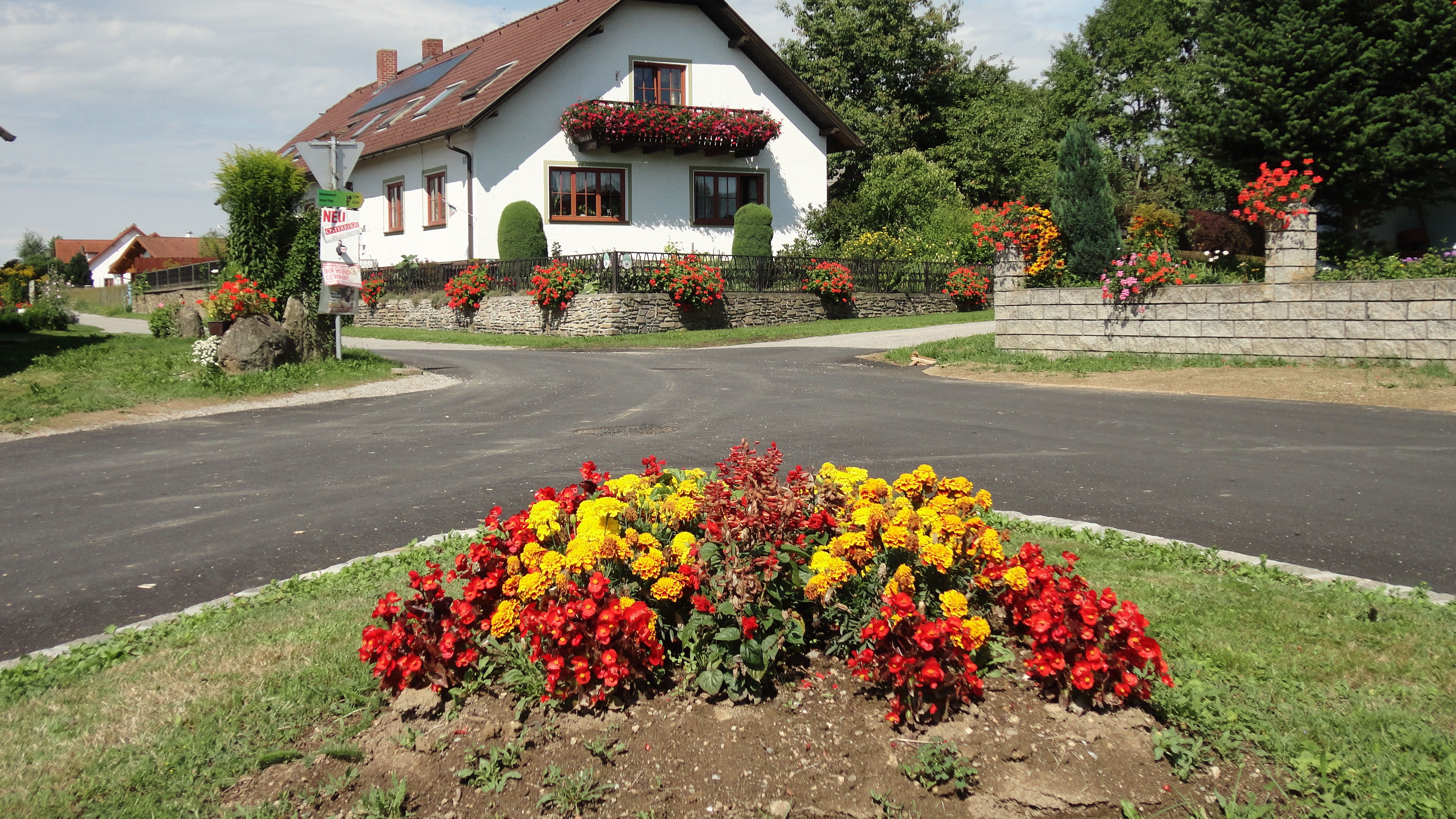 Bílý dům s červenými střešními taškami a květinovou výzdobou, obklopený barevnými záhony a zelenou krajinou.