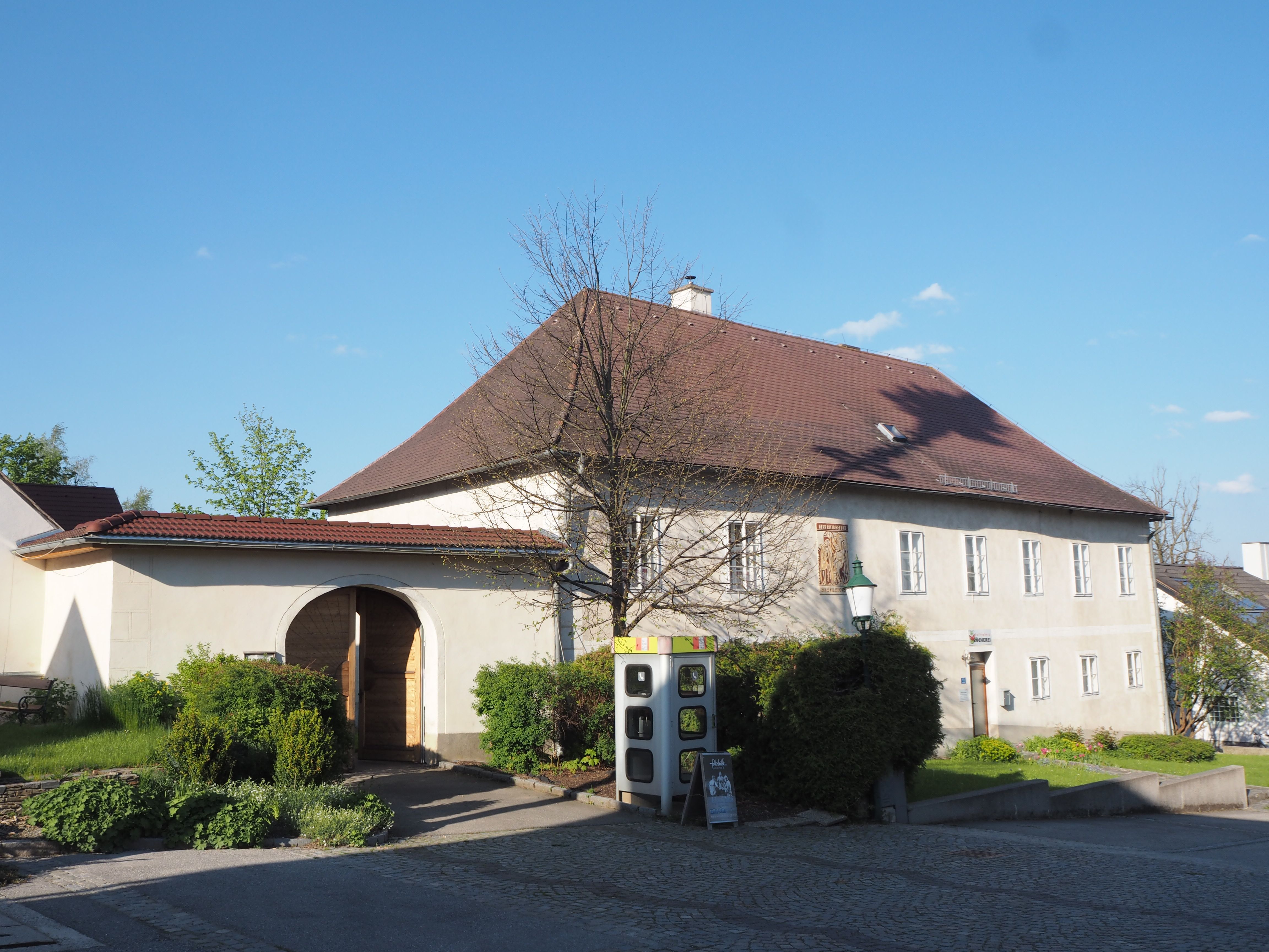 Historická budova s podloubím a zahradou, modrá obloha.