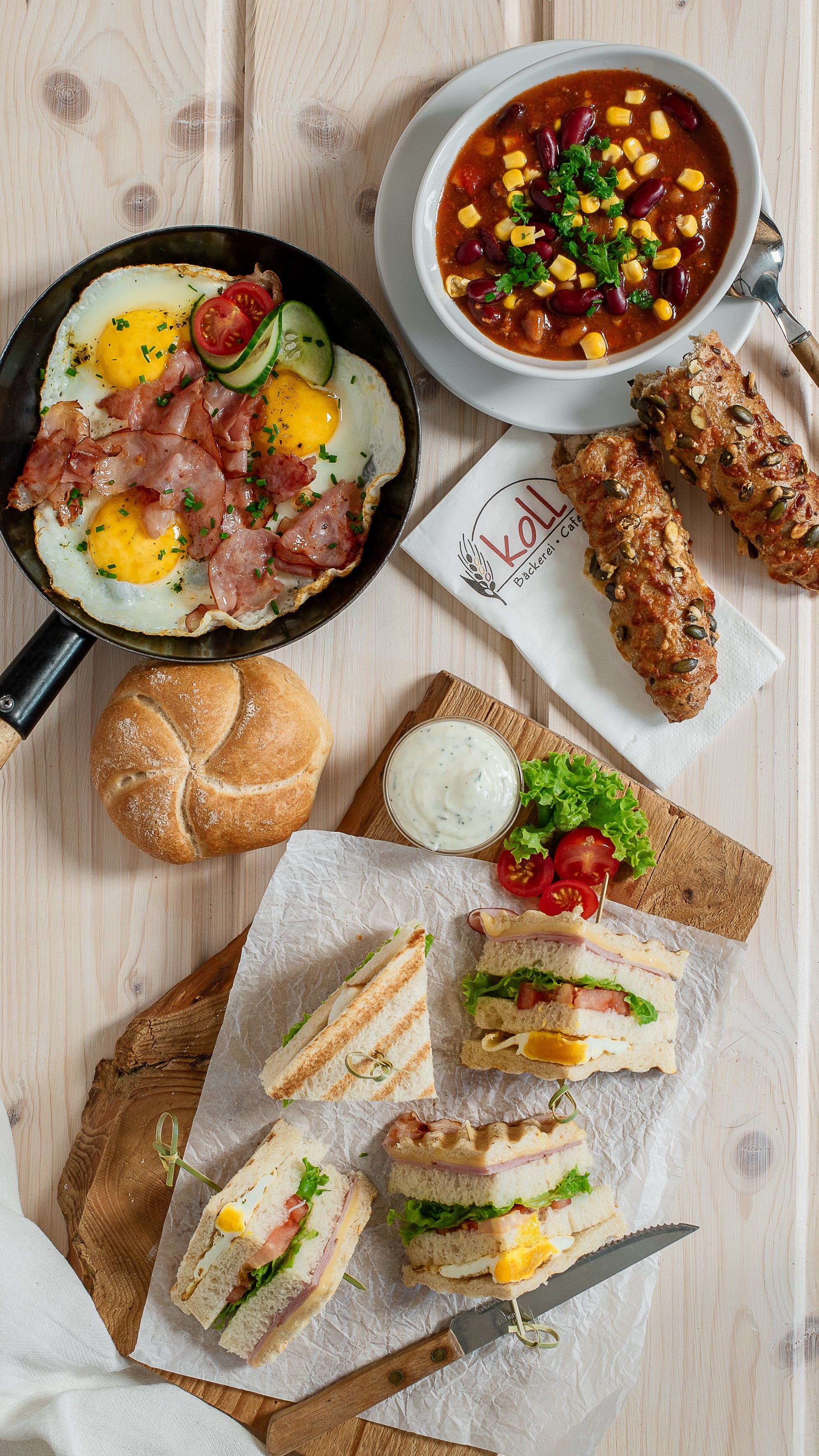 Různé teplé občerstvení na stole, včetně smažených vajec se slaninou, polévky, sendvičů a rohlíků.