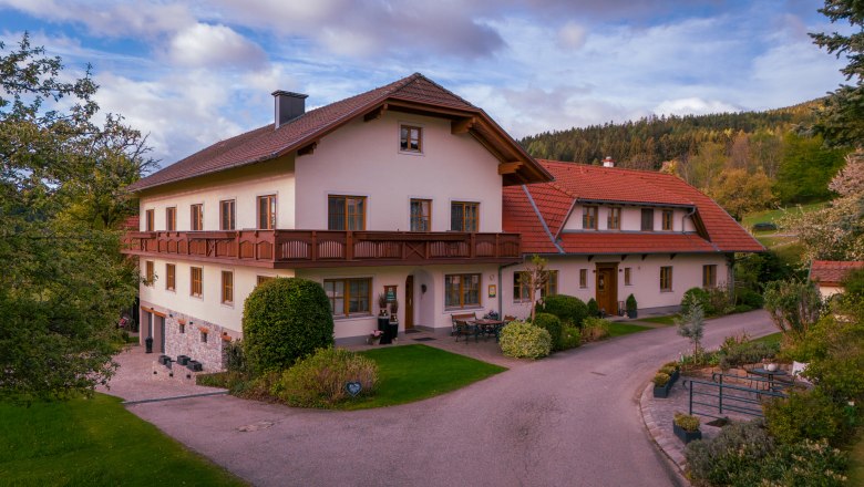 Tradiční statek s červenou střechou a balkonem, obklopený zelení a stromy.