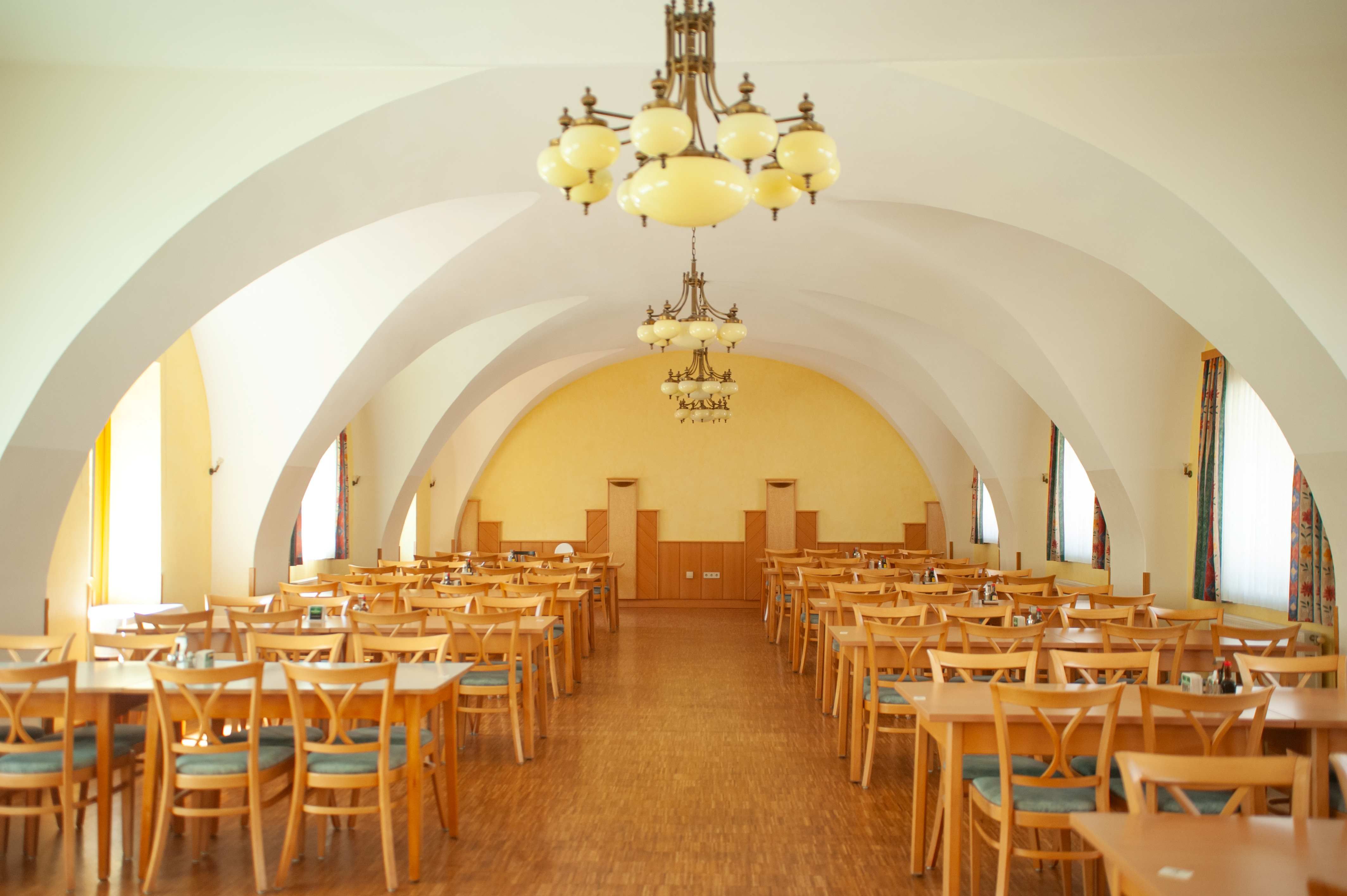 Pohled do interiéru velké jídelny s dřevěnými stoly a židlemi, žlutými stěnami a ozdobnými lustry.