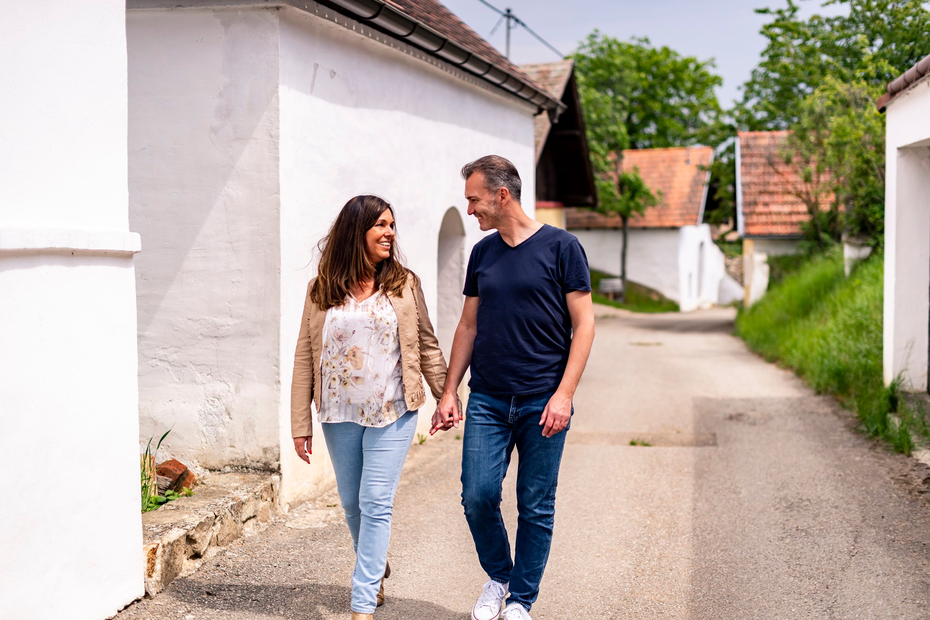 Pár se ruku v ruce prochází po Öhlbergkellergasse v Pillersdorfu, obklopen tradičními vinnými sklepy a zelenou krajinou.
