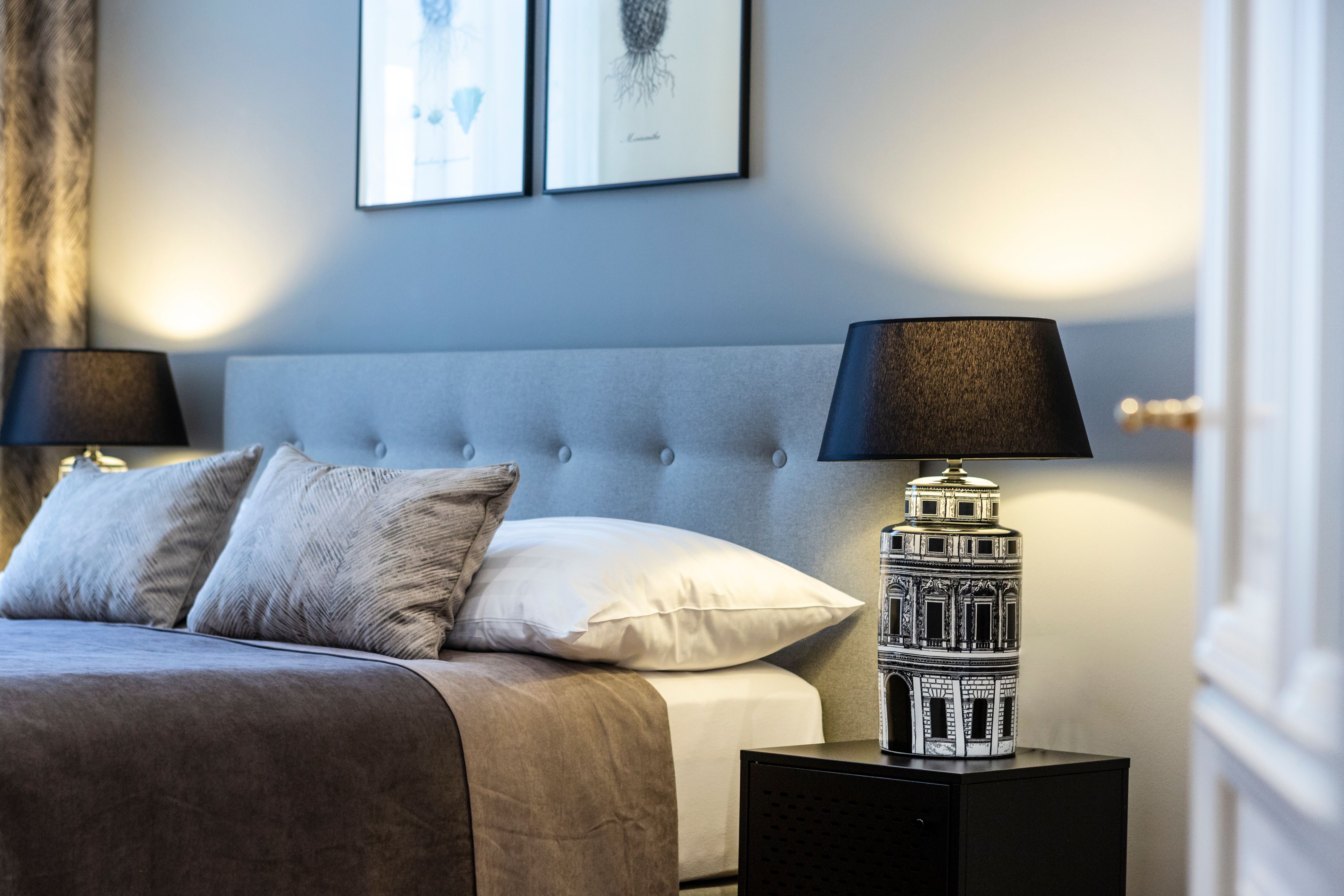 Elegantní dvoulůžkový pokoj s šedým čelem postele, dvěma polštáři, noční lampičkou a nástěnnými obrazy.