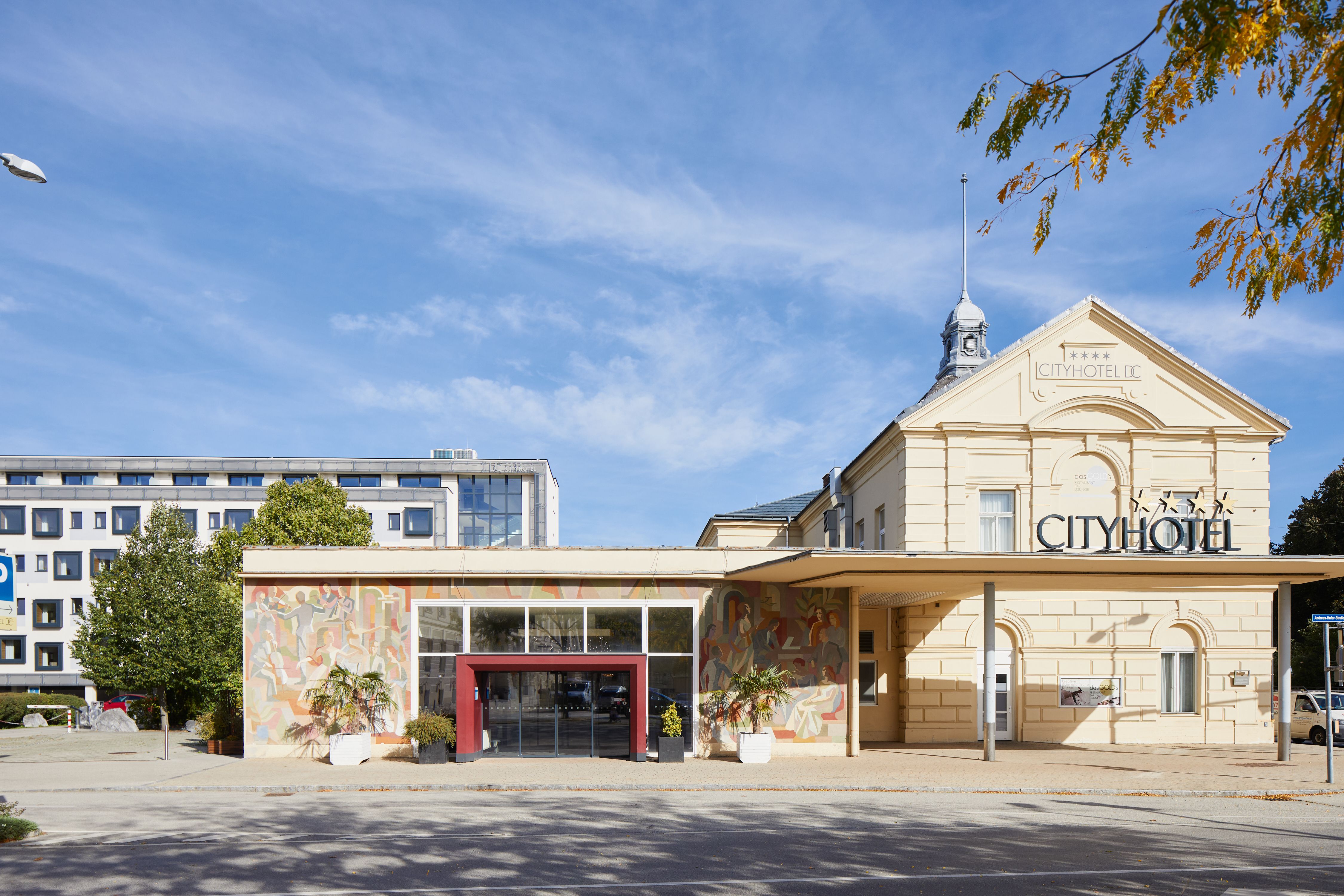 Moderní hotelová budova s prosklenými balkony a stromy v popředí.