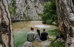 Drei Personen sitzen am Ufer der Scharzach im Höllental am Wanderweg 1. Wiener Wasserleitungsweg und machen Pause. Rundum sie ragen Felden und Bäume in die Höhe. 