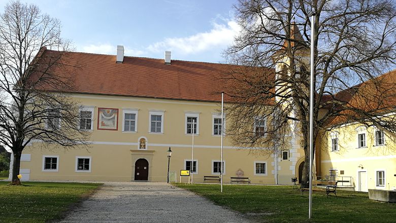 Žlutá budova hradu s červenou střechou a věží, obklopená stromy.