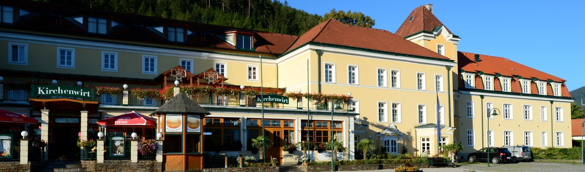 Pohled zvenčí na žlutou budovu s červenými střechami a nápisem "Kirchenwirt".