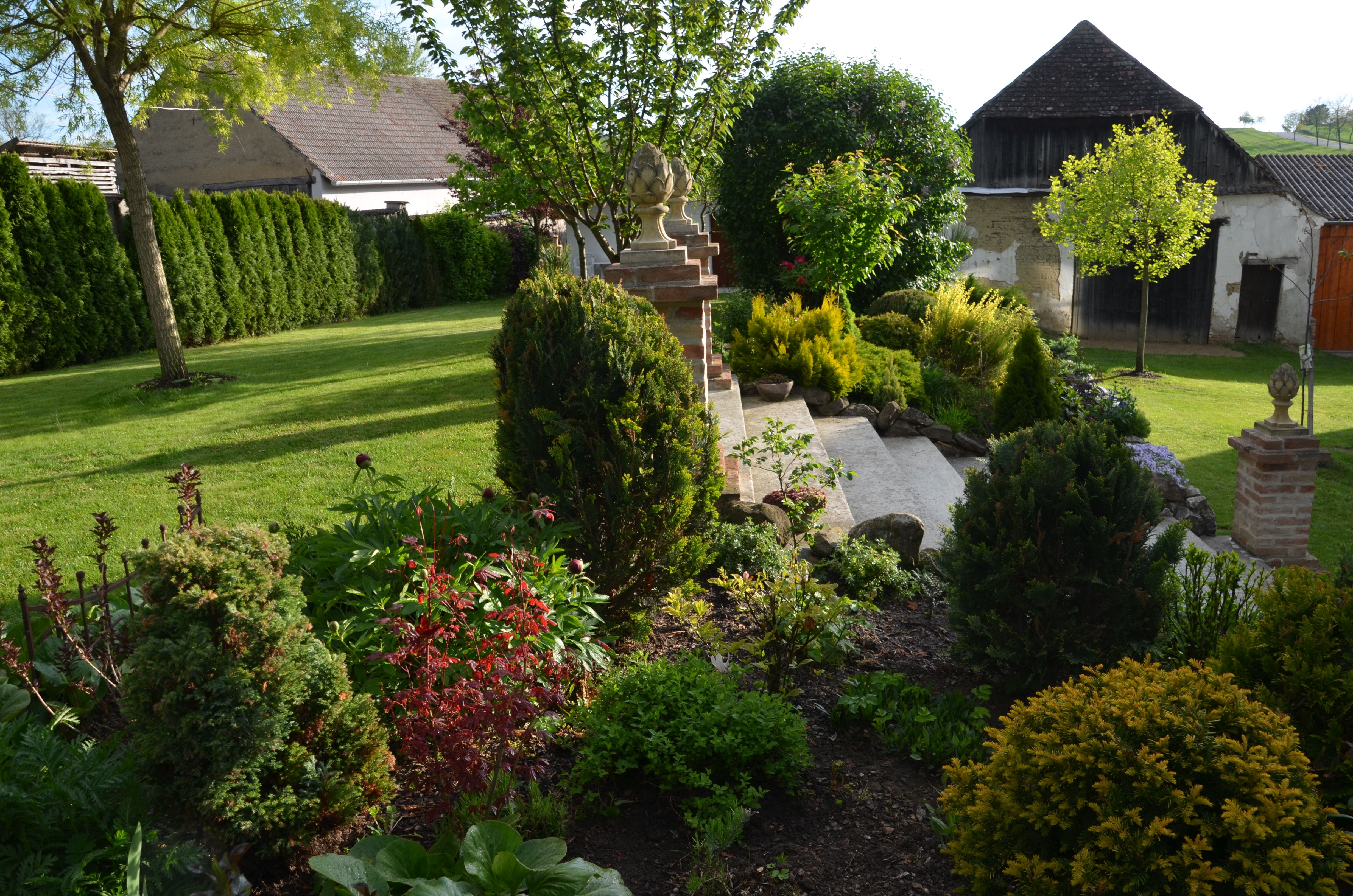 Udržovaná zahrada s trávníkem, keři a stromy, obklopená budovami v pozadí.