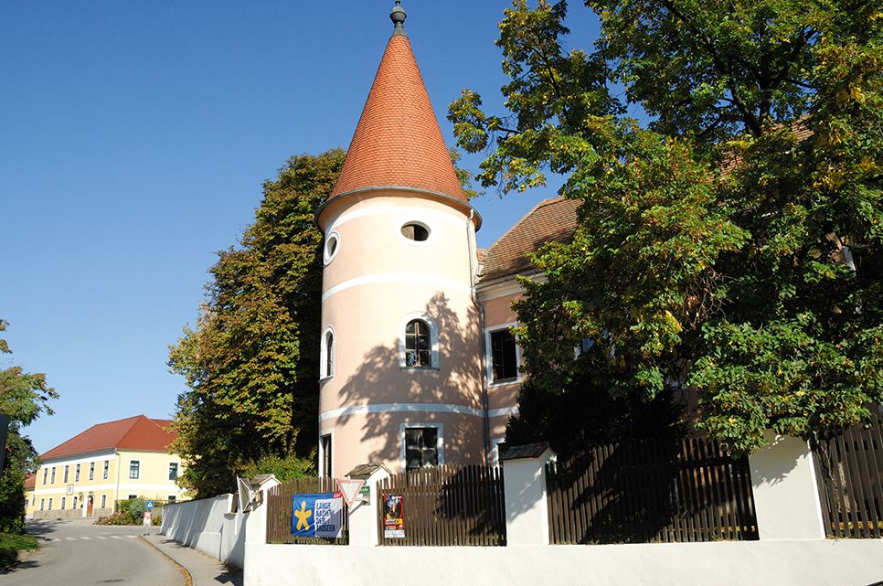 Růžová budova s věží a červenou střechou ve Fels am Wagram, obklopená stromy a modrou oblohou.