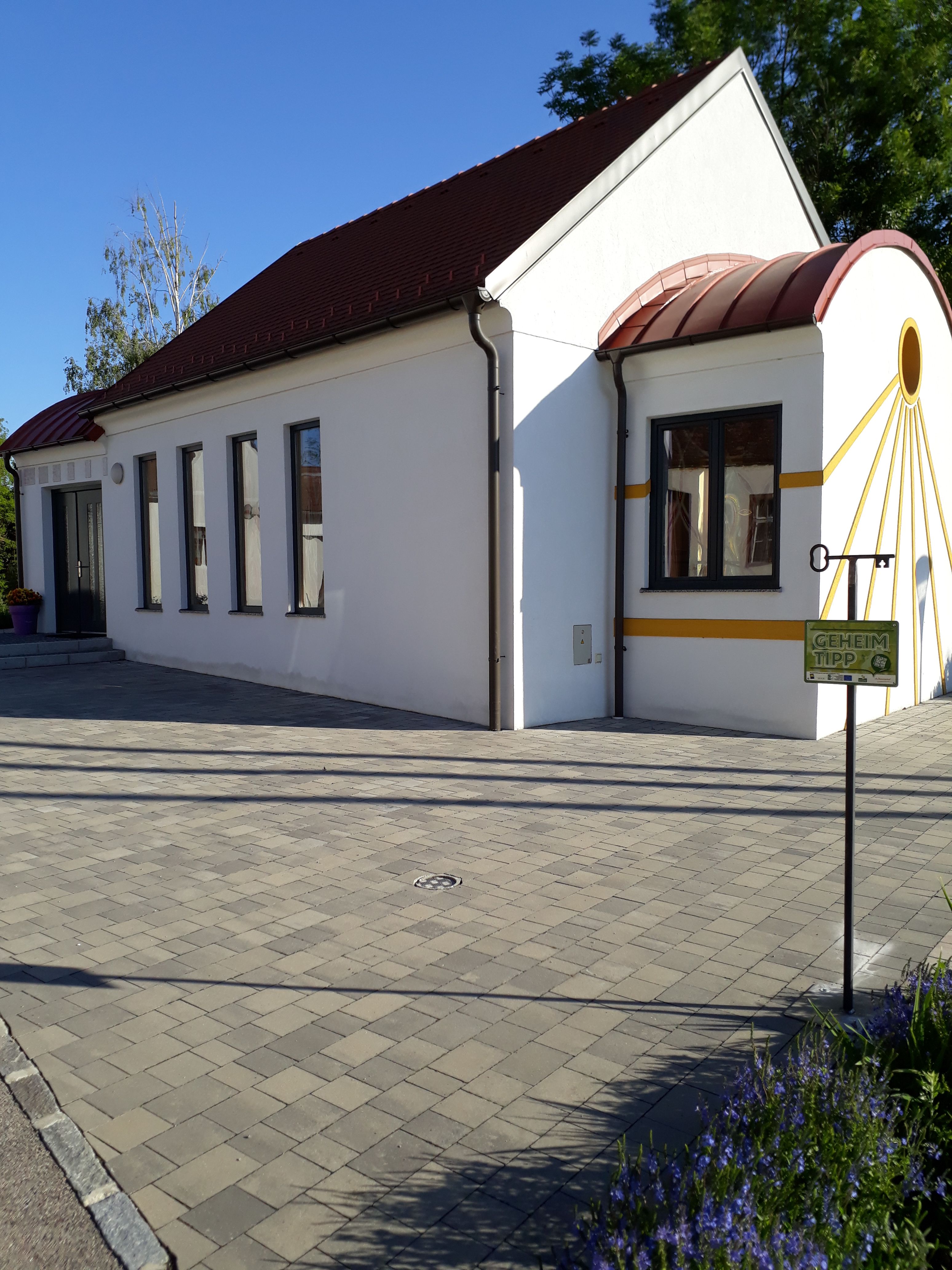 Bílá kaple s červenou střechou a žlutými akcenty, obklopená dlážděnou podlahou a rostlinami.