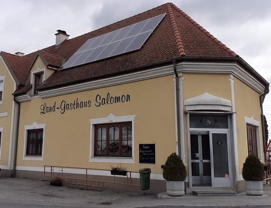 Pohled zvenčí na tradiční venkovský hostinec se světlou fasádou, červenými okenicemi a šikmou střechou.