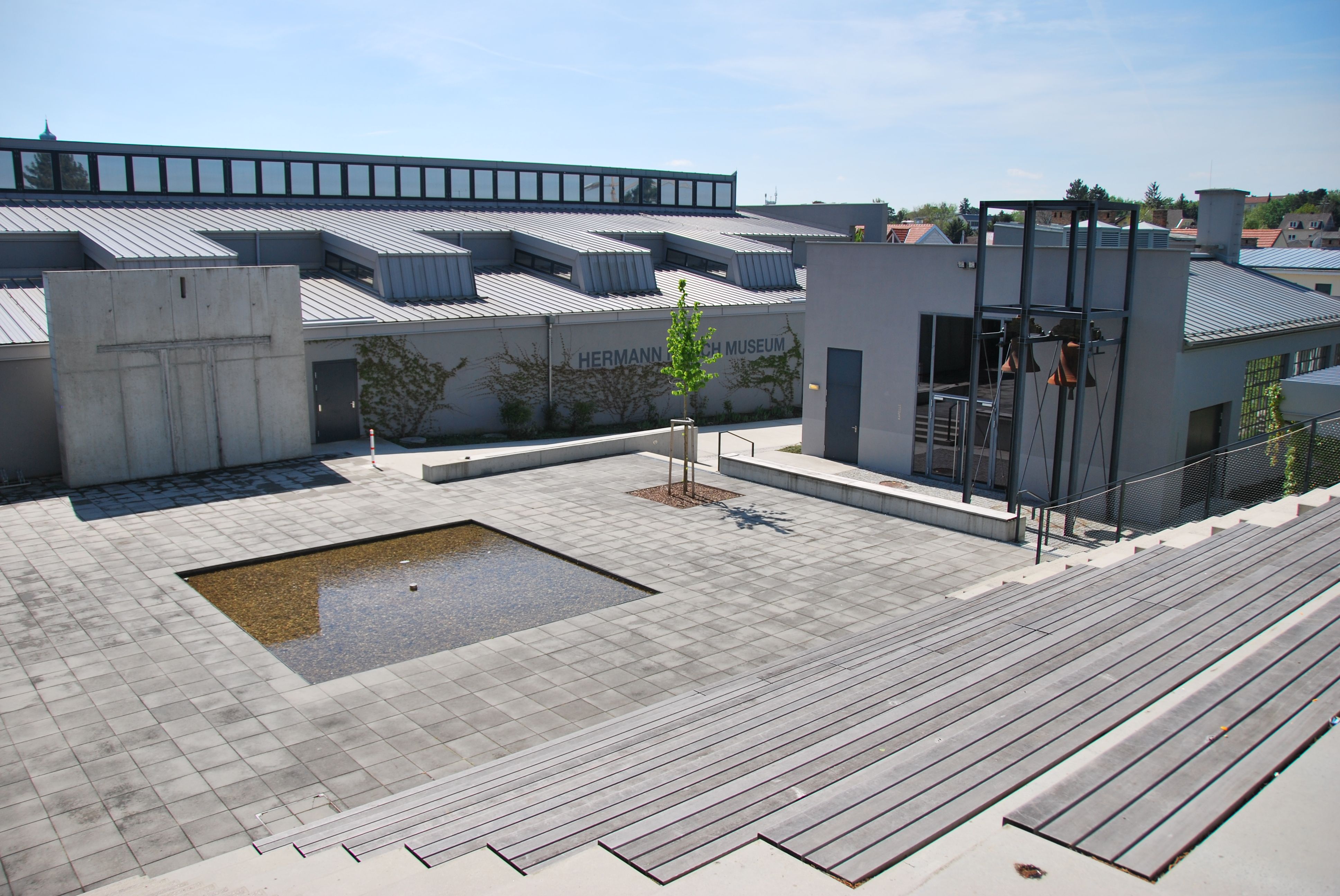 Vnější pohled na Muzeum Hermanna Nitsche s moderním designem a malou vodní nádrží na náměstí.