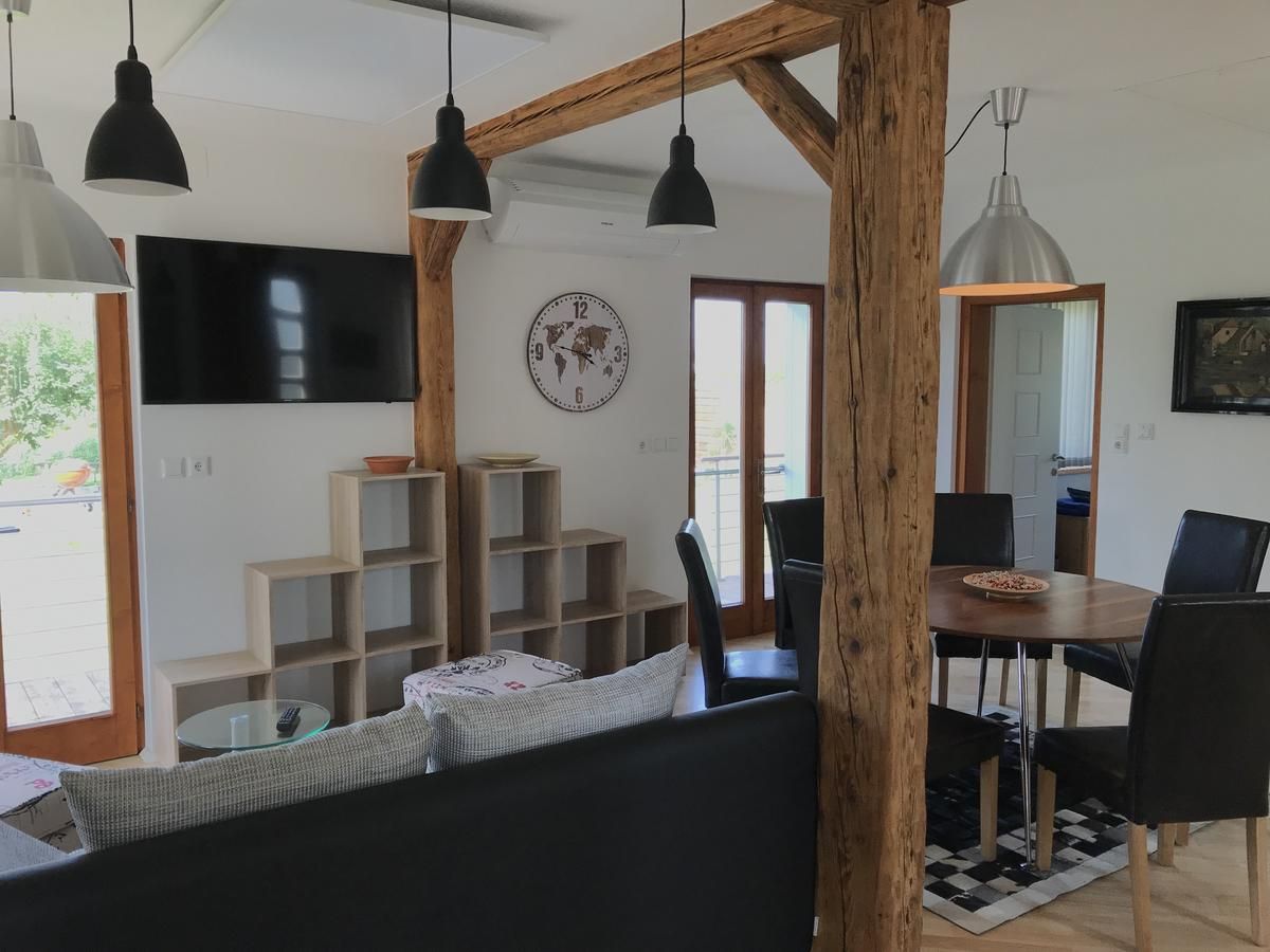 Moderní obývací prostor s dřevěnými trámy, jídelním stolem, židlemi, nástěnnou policí, hodinami a TV.