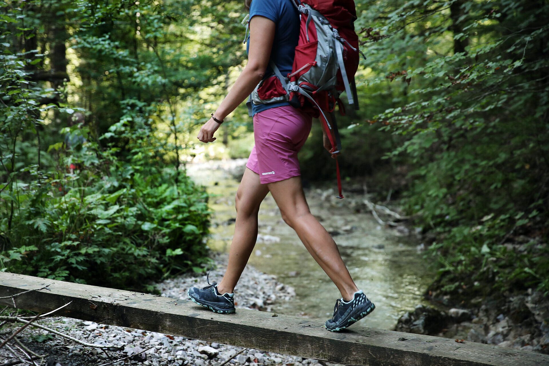 Žena v turistickém oblečení balancuje na dřevěných kůlech nad malým potokem. 