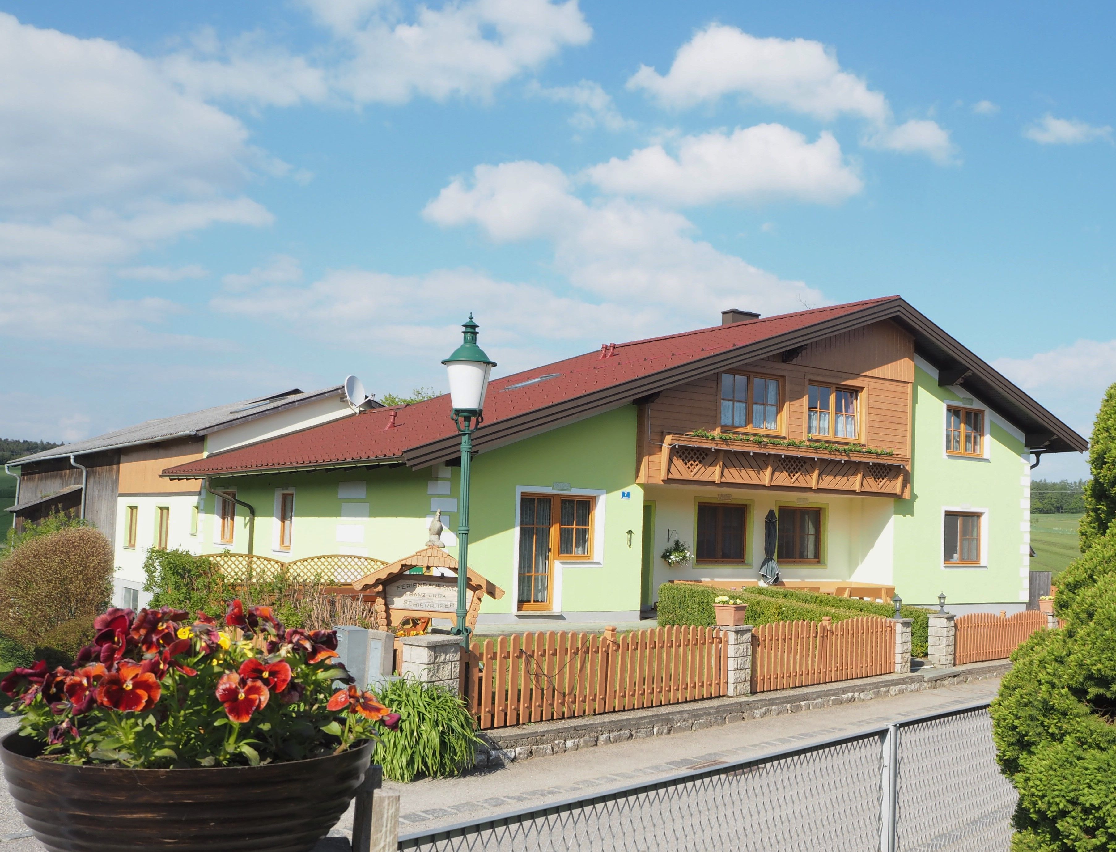 Rekreační statek se zeleným nátěrem a dřevěným balkonem, obklopený dřevěným plotem a květinami v popředí.