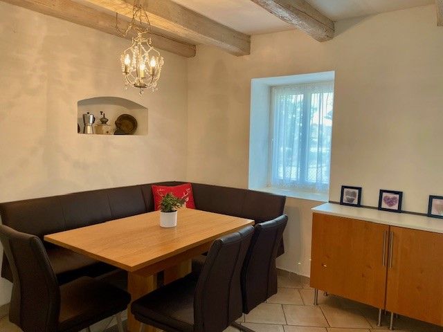 Útulný jídelní kout s dřevěným stolem, černými židlemi a rohovou lavicí. Na stěně visí lustr, vedle něj okno a příborník.