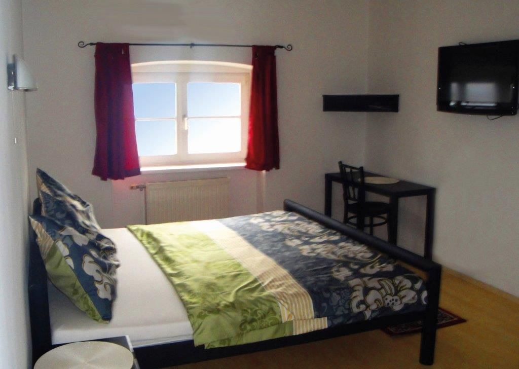 Ložnice s postelí, oknem s červenými závěsy, malým stolkem a židlí a nástěnnou televizí.