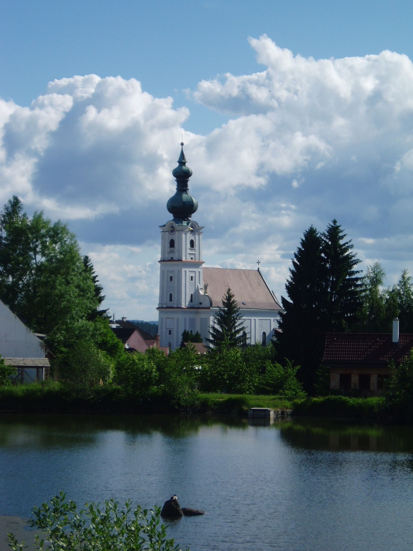 Kostel v Kirchbergu am Walde s rybníkem v popředí a zataženou oblohou.