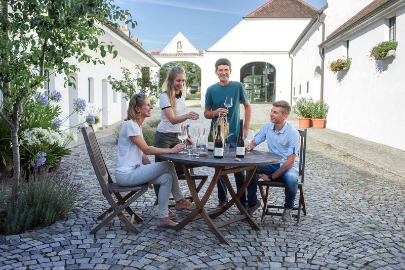 Čtyři lidé sedí venku u kulatého stolu a vychutnávají si víno. Jsou obklopeni bílými budovami.
