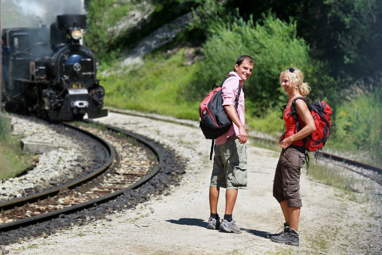Dva turisté stojí na štěrkové cestě vedle železničních kolejí a kolem projíždí parní lokomotiva.