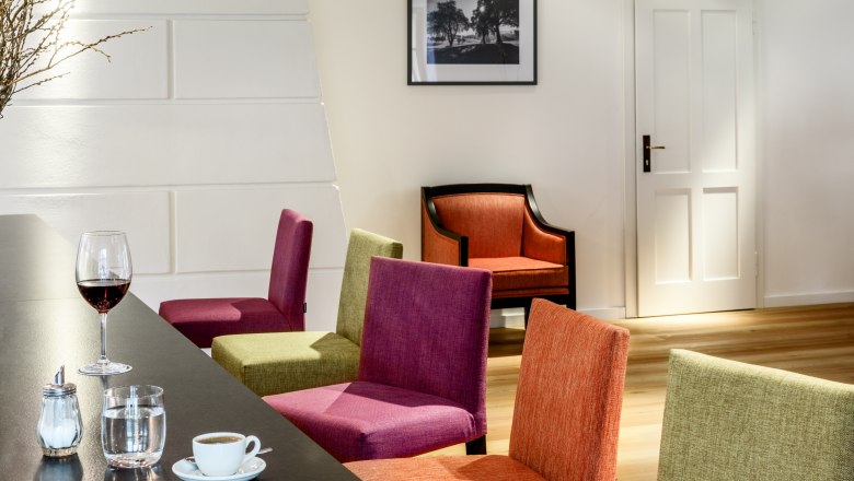 Pohled do interiéru moderního hotelového salónku s barevnými židlemi, stolkem s nápoji a obrazem na stěně.