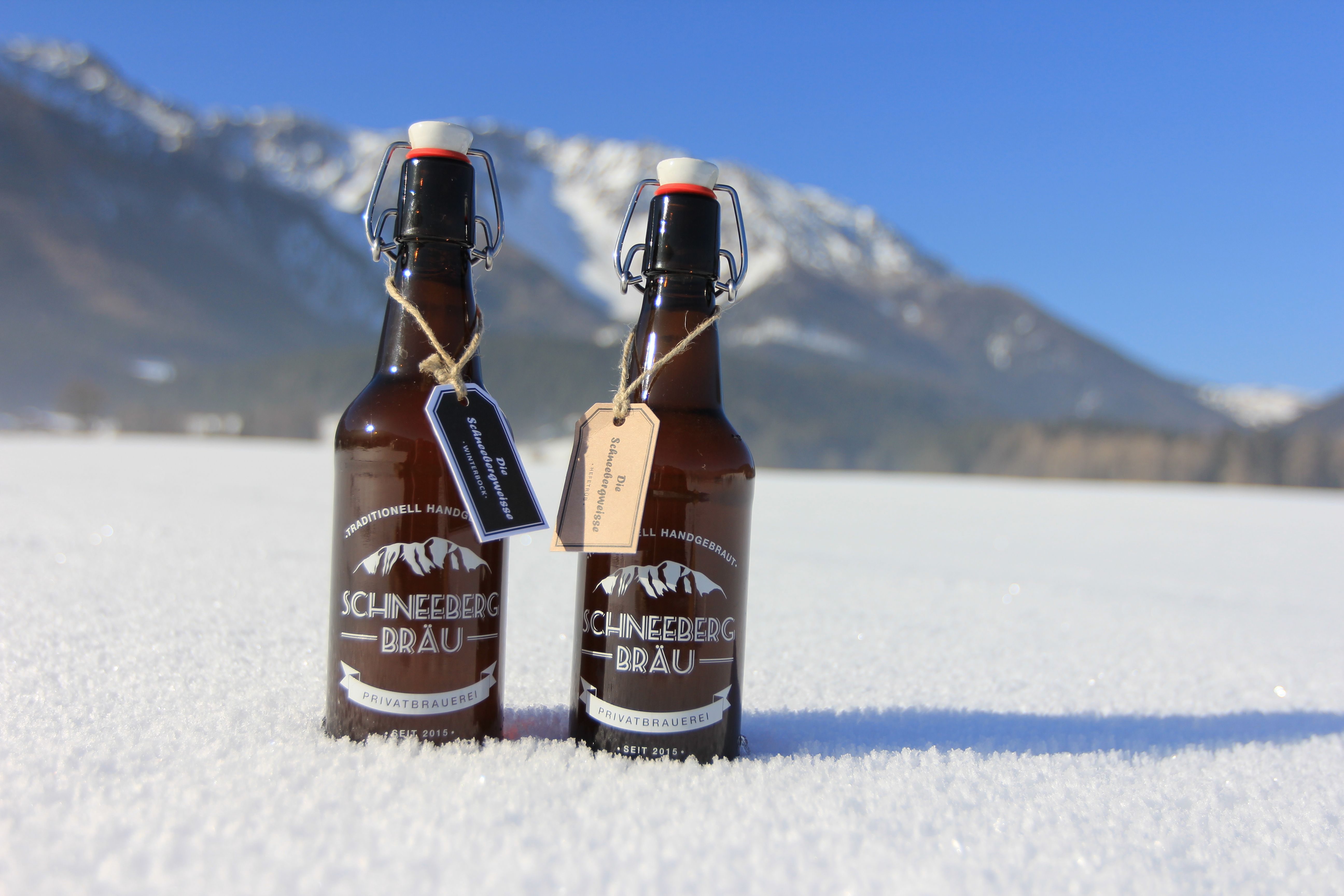 Dvě lahve piva Schneeberg Bräu stojí na sněhu na pozadí hor.