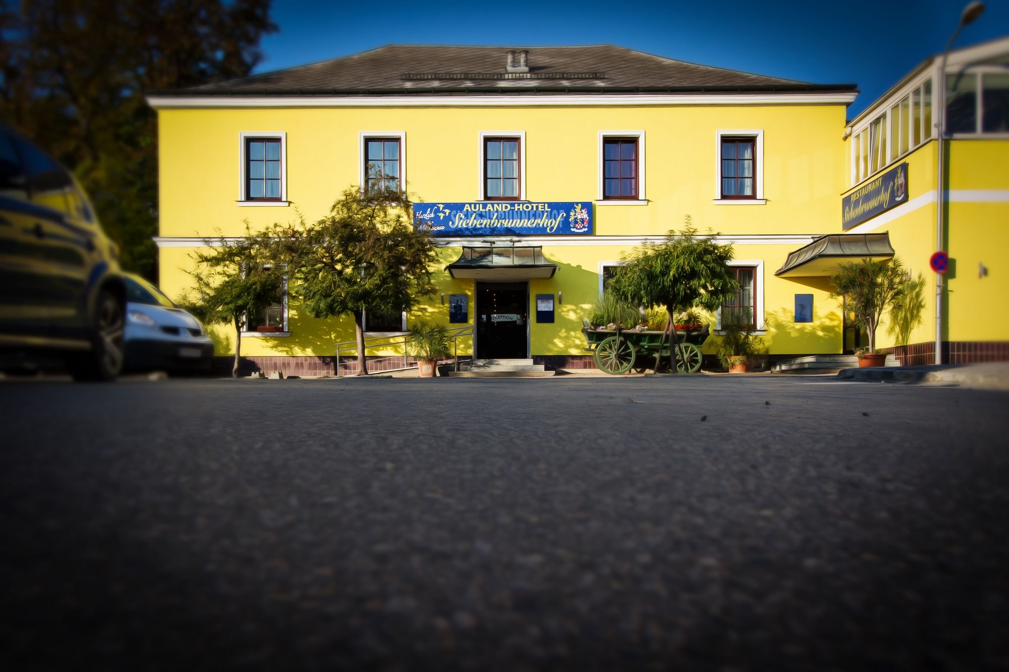 Žlutá budova s nápisem "Auland-Hotel Siebenbrunnerhof", obklopená stromy a starým kočárem.