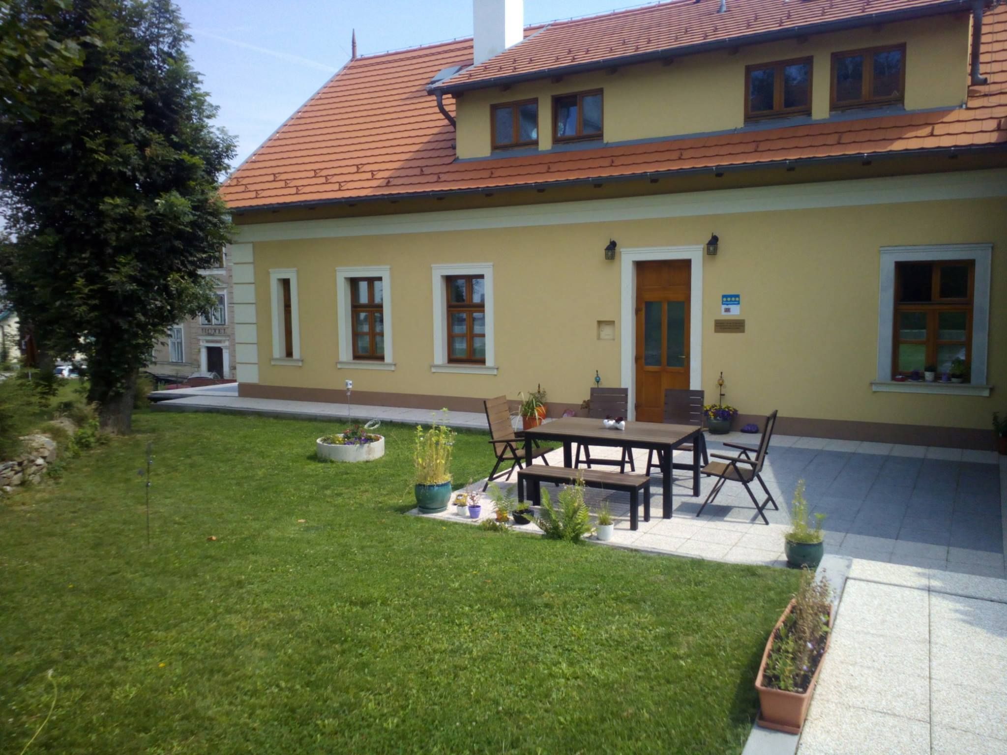 Žlutý dům s červenou střechou a terasou, obklopený trávníkem a rostlinami.