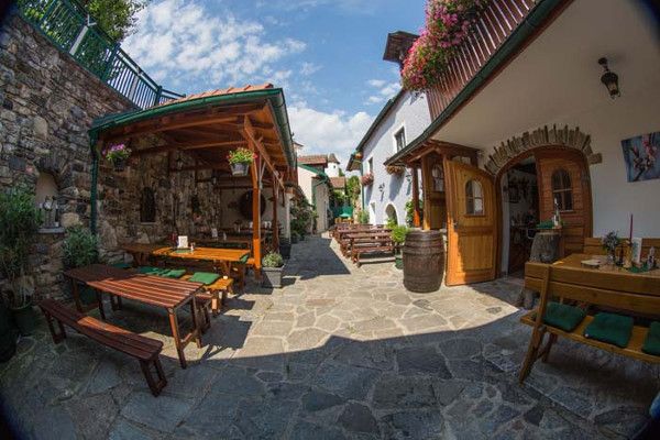 Útulný dvůr s dřevěnými lavicemi, stoly a květinami. Kamenná podlaha a kryté posezení. Slunečné počasí s modrou oblohou.