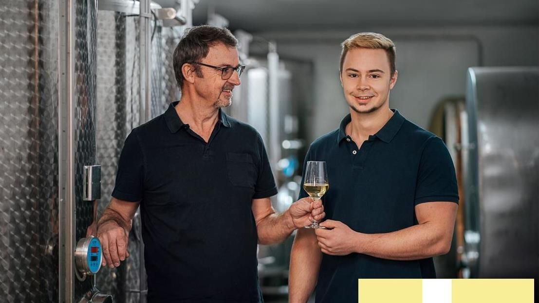 Dva muži ve vinném sklípku, jeden drží sklenici bílého vína, druhý podává cisternu.