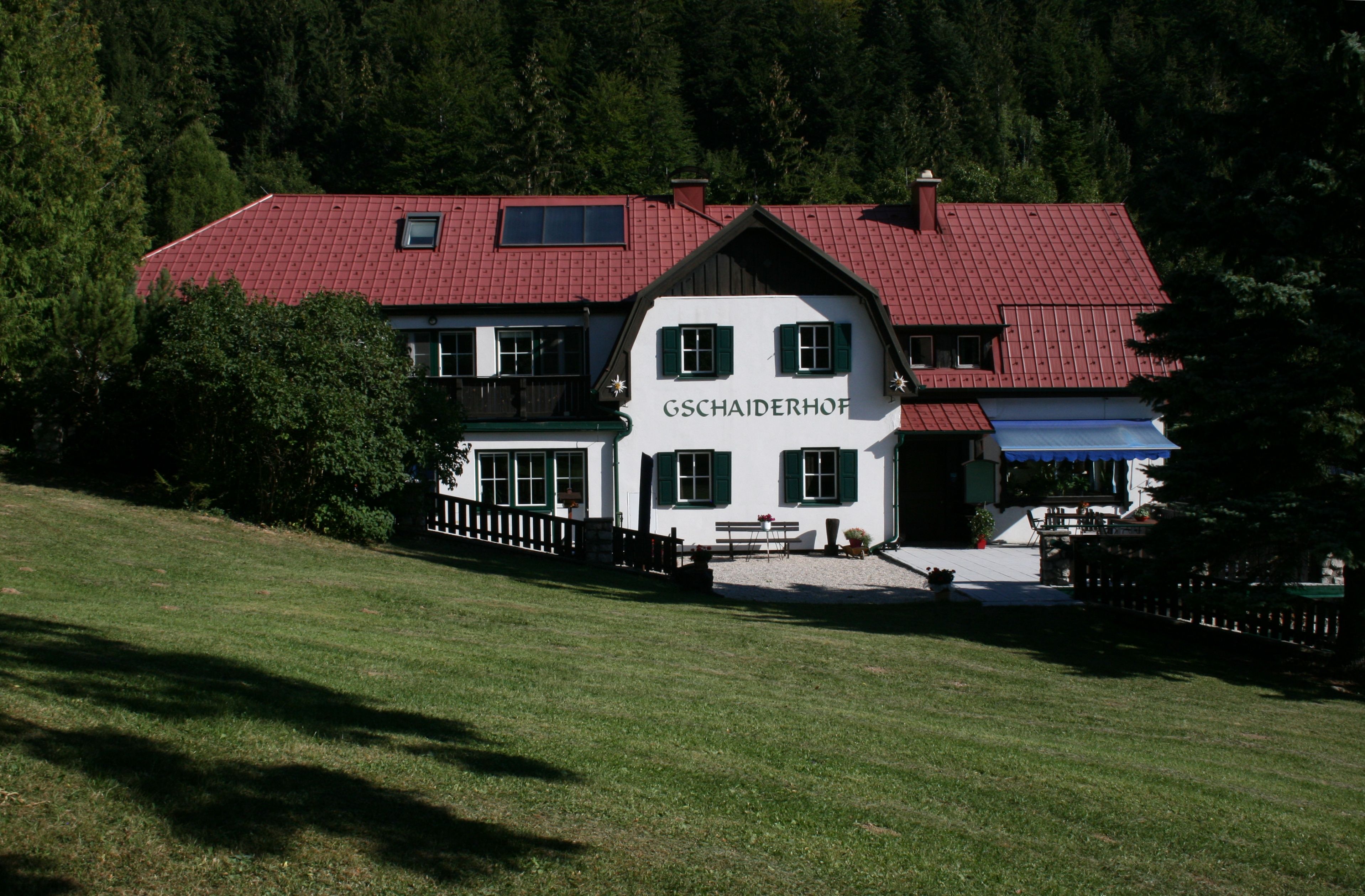 Tradiční budova s červenou střechou a zelenými okenicemi, obklopená stromy a loukami, s nápisem "Gschaiderhof".