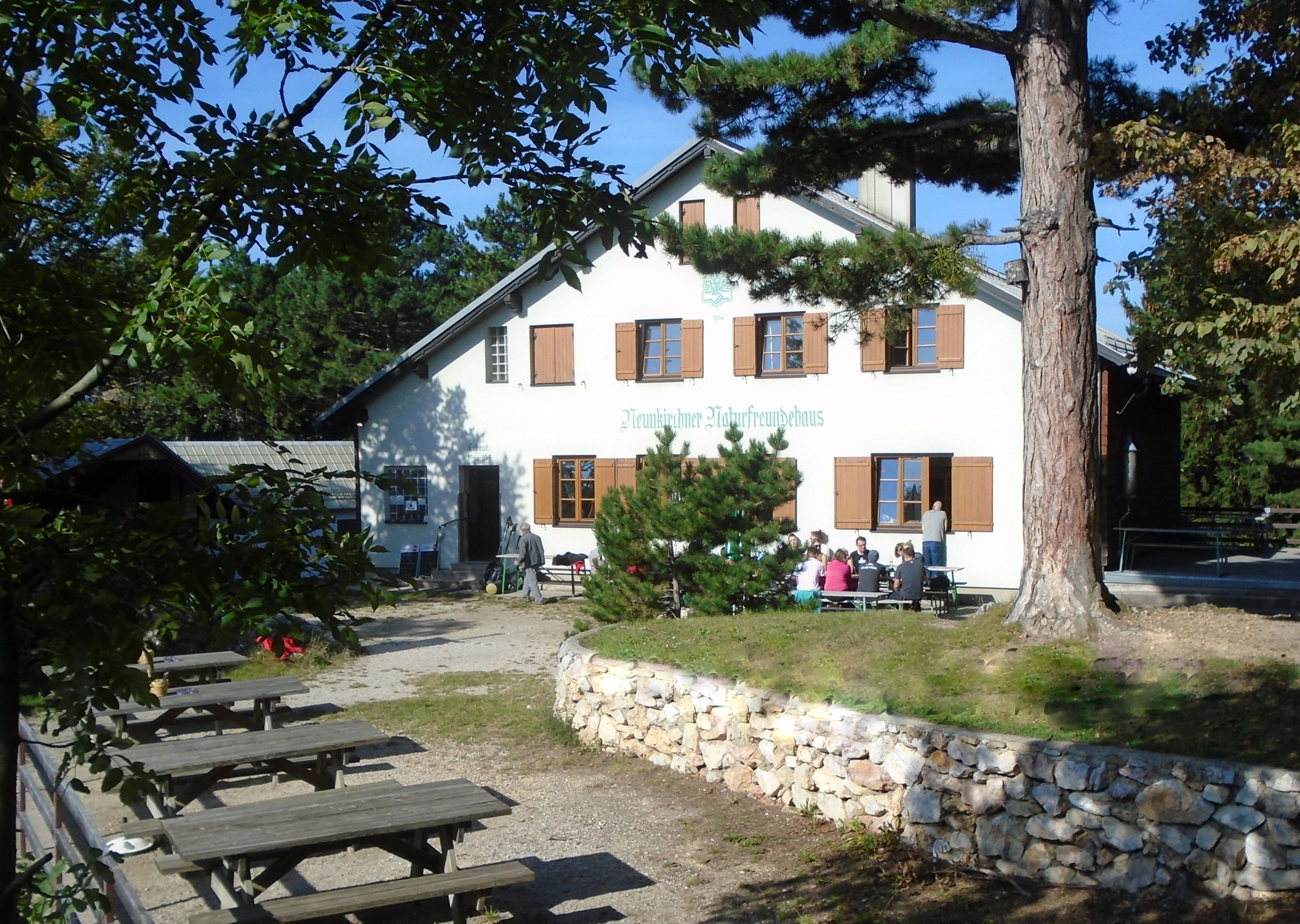 Budova s nápisem "Neunkirchner Naturfreundehaus", obklopená stromy a lavičkami.