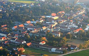 Letecký pohled na vesnici s kostelem a domy, obklopenou stromy a poli.