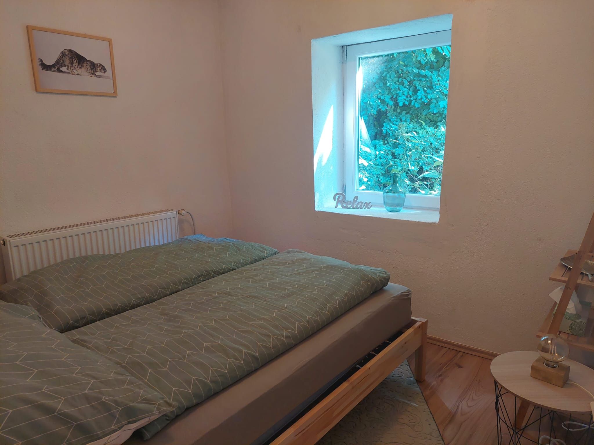 Útulná ložnice s postelí, oknem s výhledem do zeleně a obrazem na stěně.