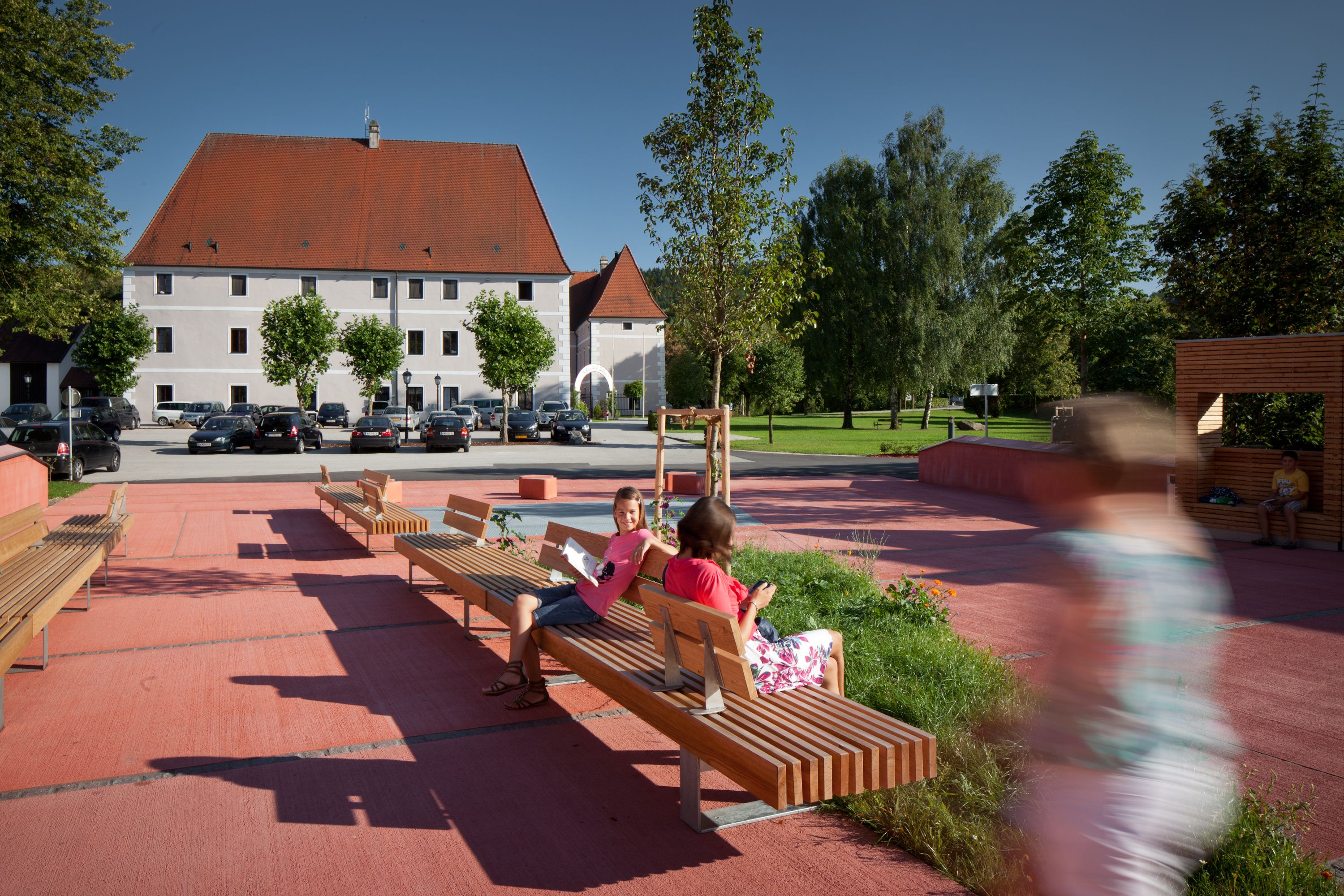 Moderní náměstí s dřevěnými lavičkami a lidmi ve slunném prostředí, s velkou budovou s červenou střechou v pozadí.