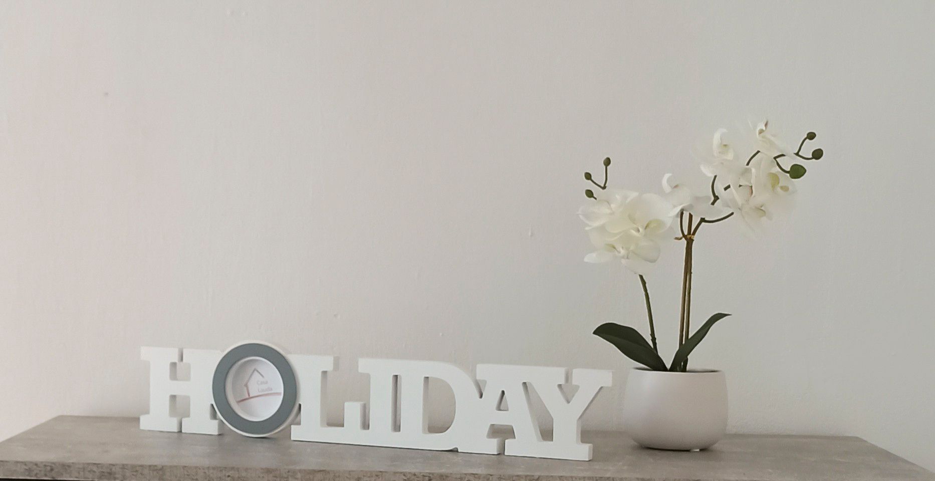 Dekorace s nápisem "HOLIDAY" a bílou orchidejí v květináči na stole.