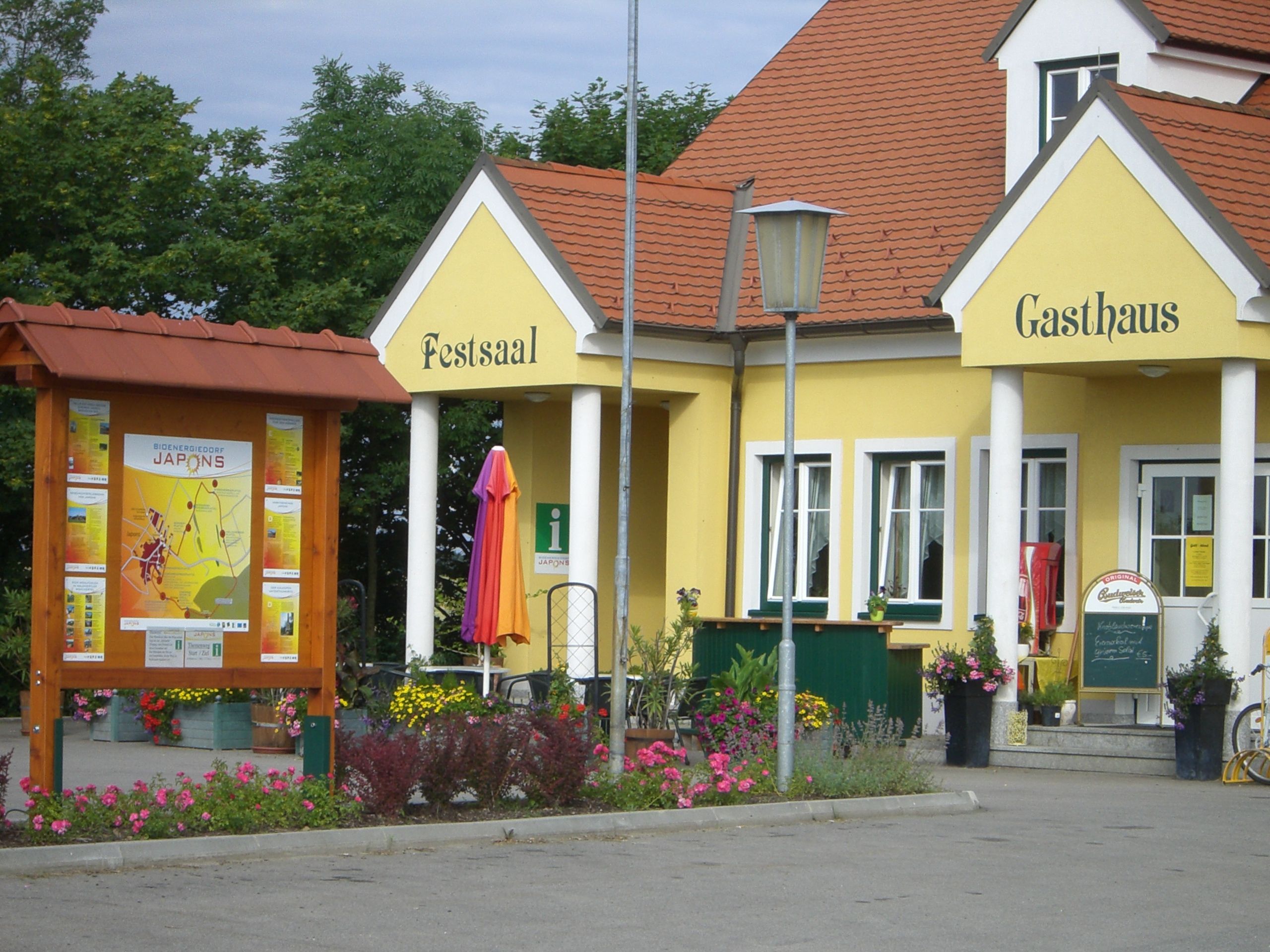 Žlutá budova s nápisy "Festsaal" a "Gasthaus", obklopená květinami a informační cedulí.