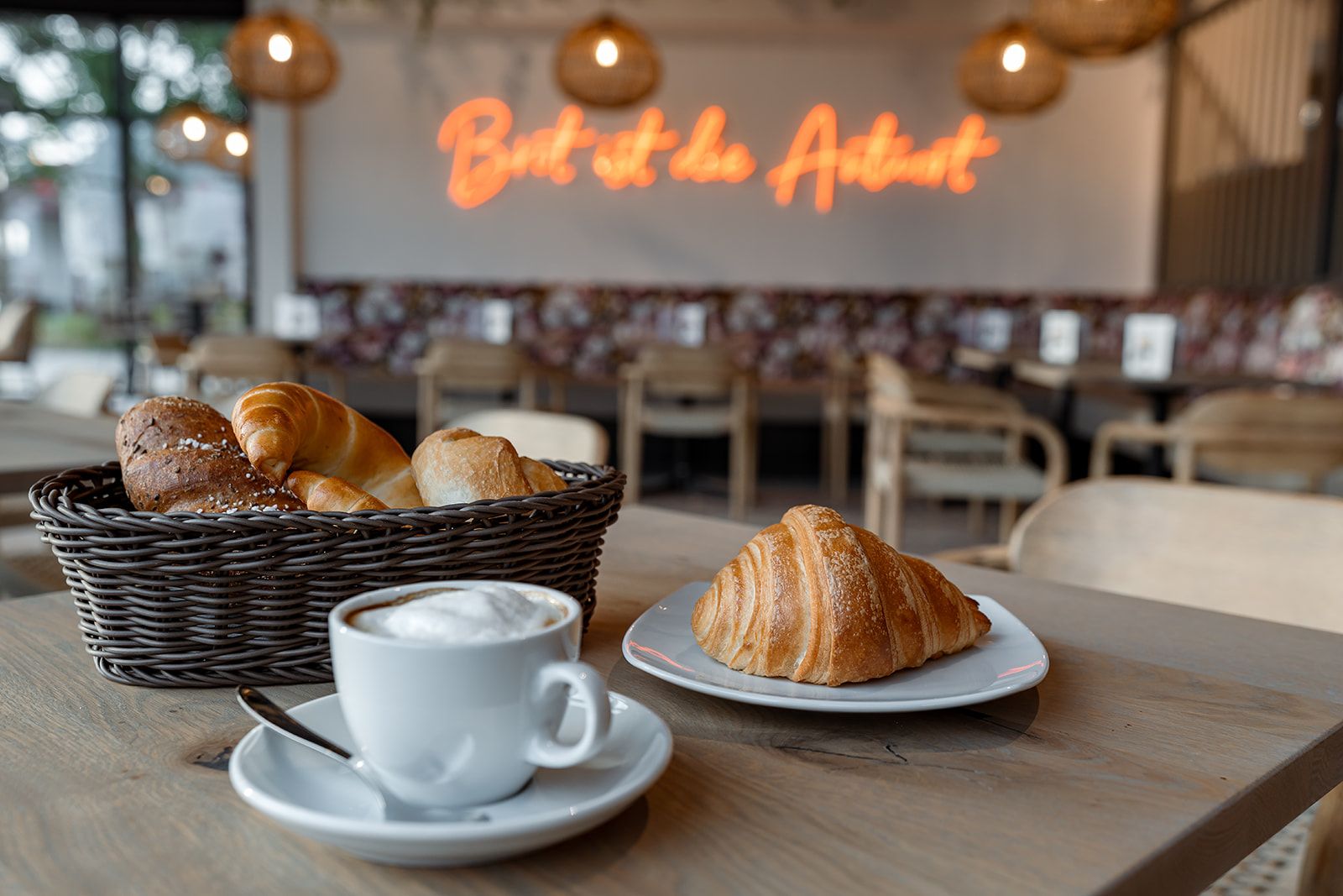 Kavárna s košíkem pečiva, croissantem na talíři a šálkem cappuccina na stole.