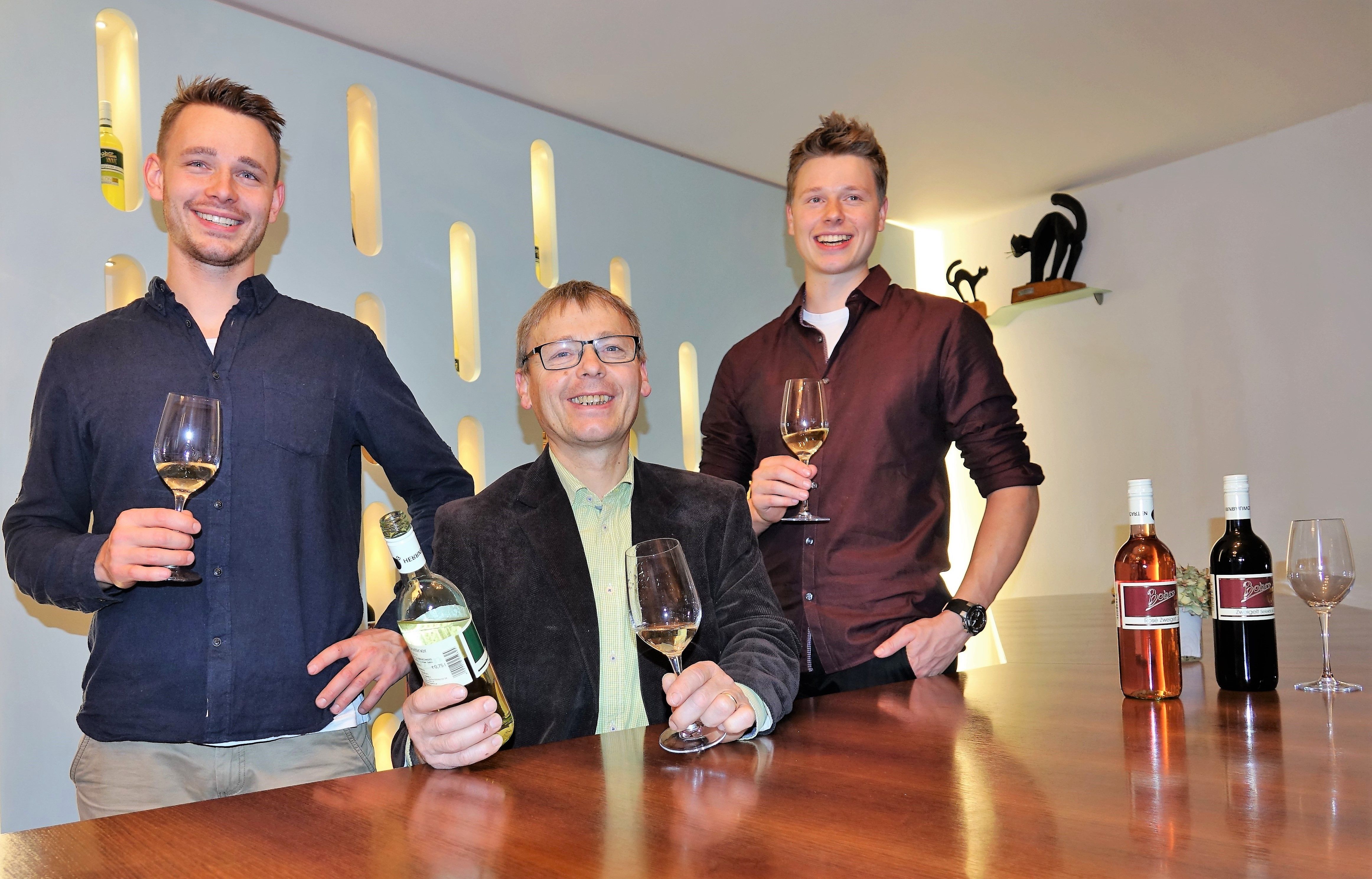 Tři muži stojí u stolu se sklenicemi a lahvemi vína.
