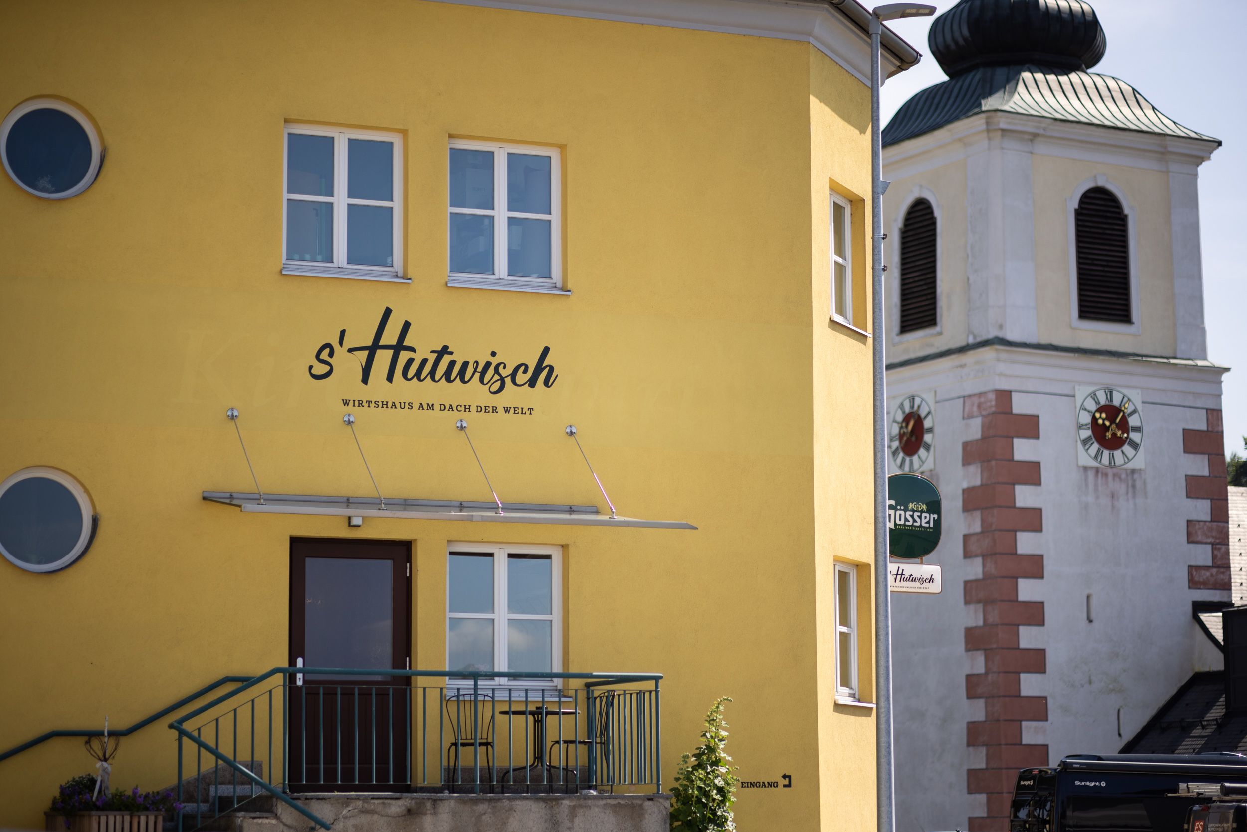 Žlutá budova s nápisem "s'Hutwisch" vedle kostelní věže s hodinami.