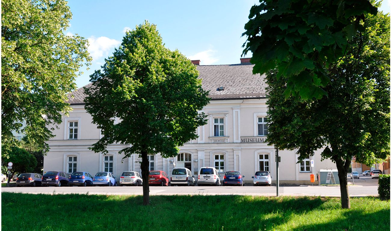 Bílá budova s nápisem "Muzeum", obklopená stromy a zaparkovanými auty.
