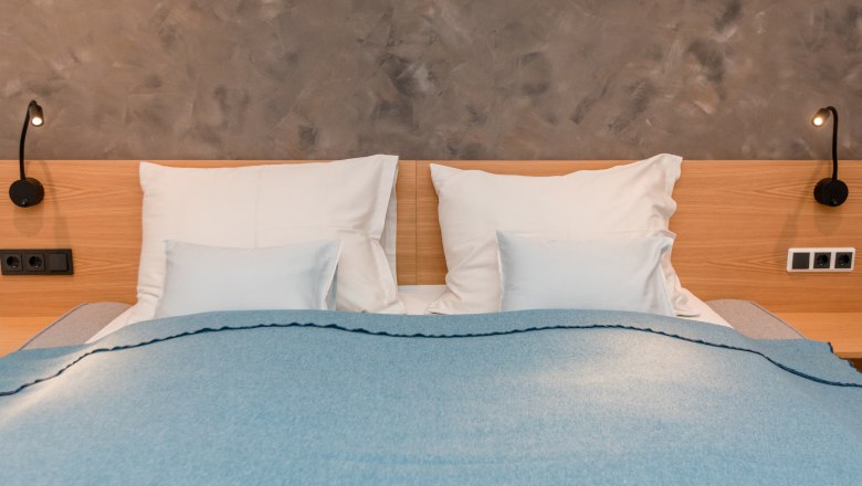 Manželská postel s modrou dekou a bílými polštáři před šedou stěnou.