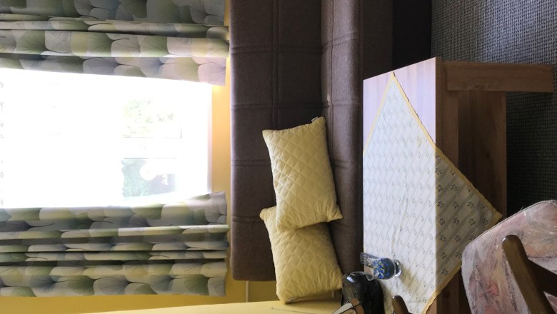 Ob&yacute;vac&iacute; pokoj s hnědou pohovkou, žlut&yacute;mi pol&scaron;t&aacute;ři, stolem s ubrusem, křeslem a oknem se vzorovan&yacute;mi z&aacute;věsy.